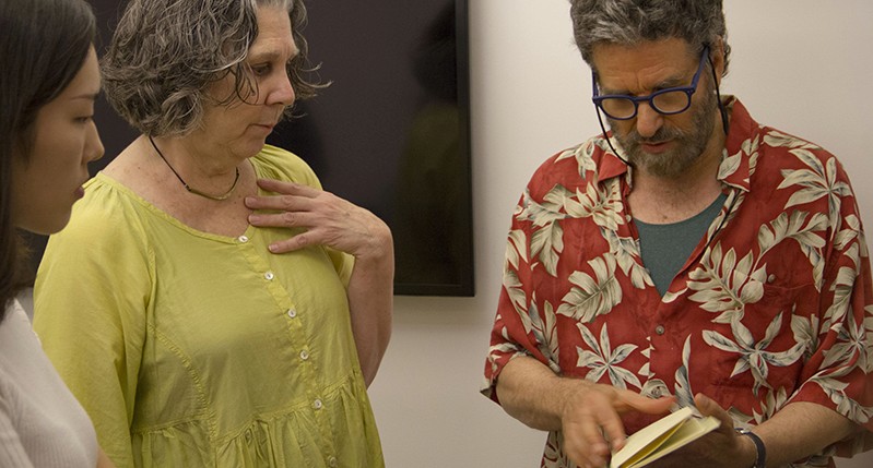 A photo of a man reading something from a flyer while two women are listening and watching.