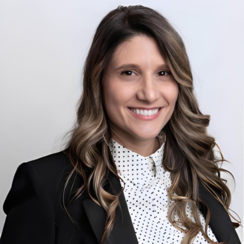 A man with glasses and a light gray blazer smiles against a softly lit background.