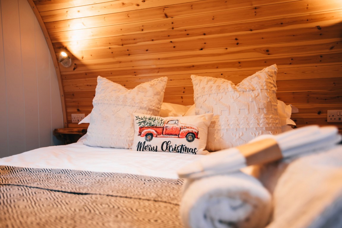 A bed with a christmas cushion on it. Slippers and a towel are in the foreground.