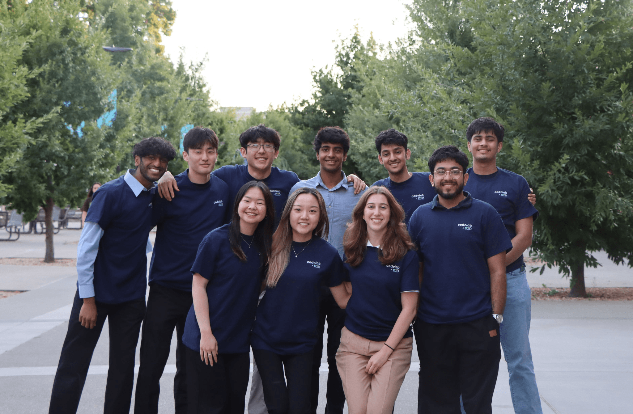 10 students posing together with matching shirts, green trees and bright sky in the background