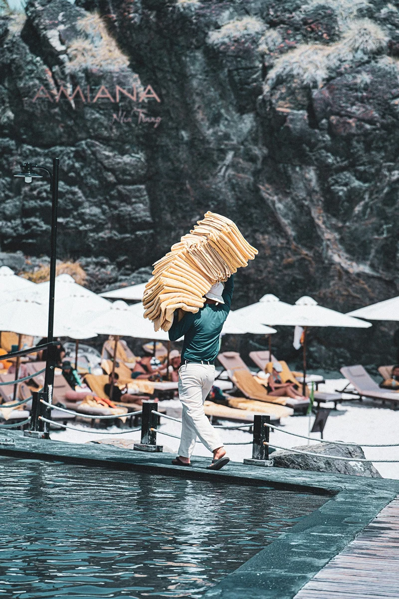 Resort staff carrying stacked lounge cushions near an outdoor pool area.