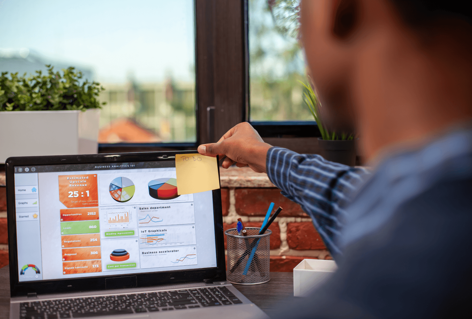 A marketing professional analyzing a complex business analytics dashboard on a laptop, featuring charts for revenue, lead opportunities, and sales department metrics while placing a yellow sticky note on the screen.