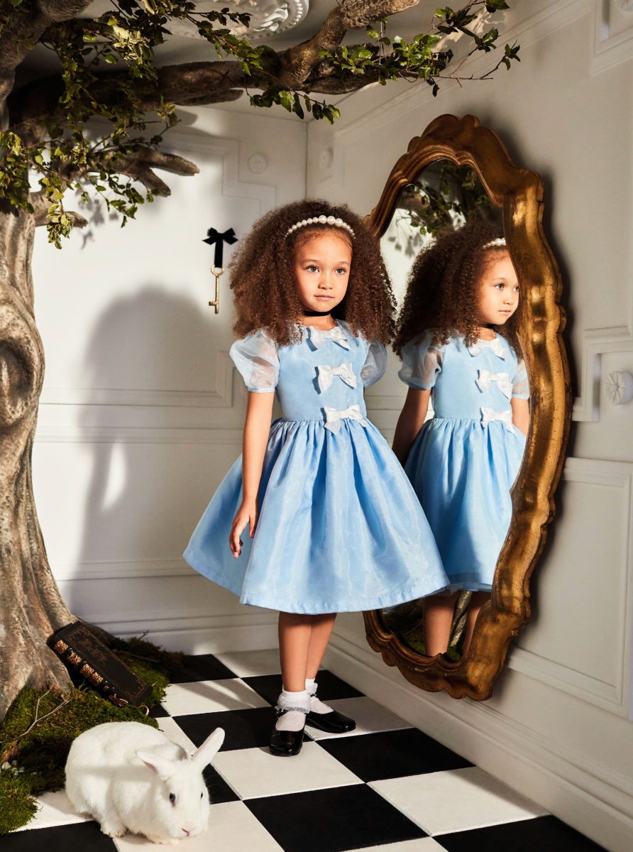 A girl in a blue dress stands in front of a mirror, with a checkered floor and whimsical decor around her.
