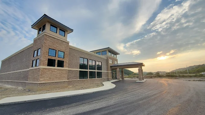 A modern building with a tower, large windows, and a paved entrance, set against a cloudy sky at sunset.