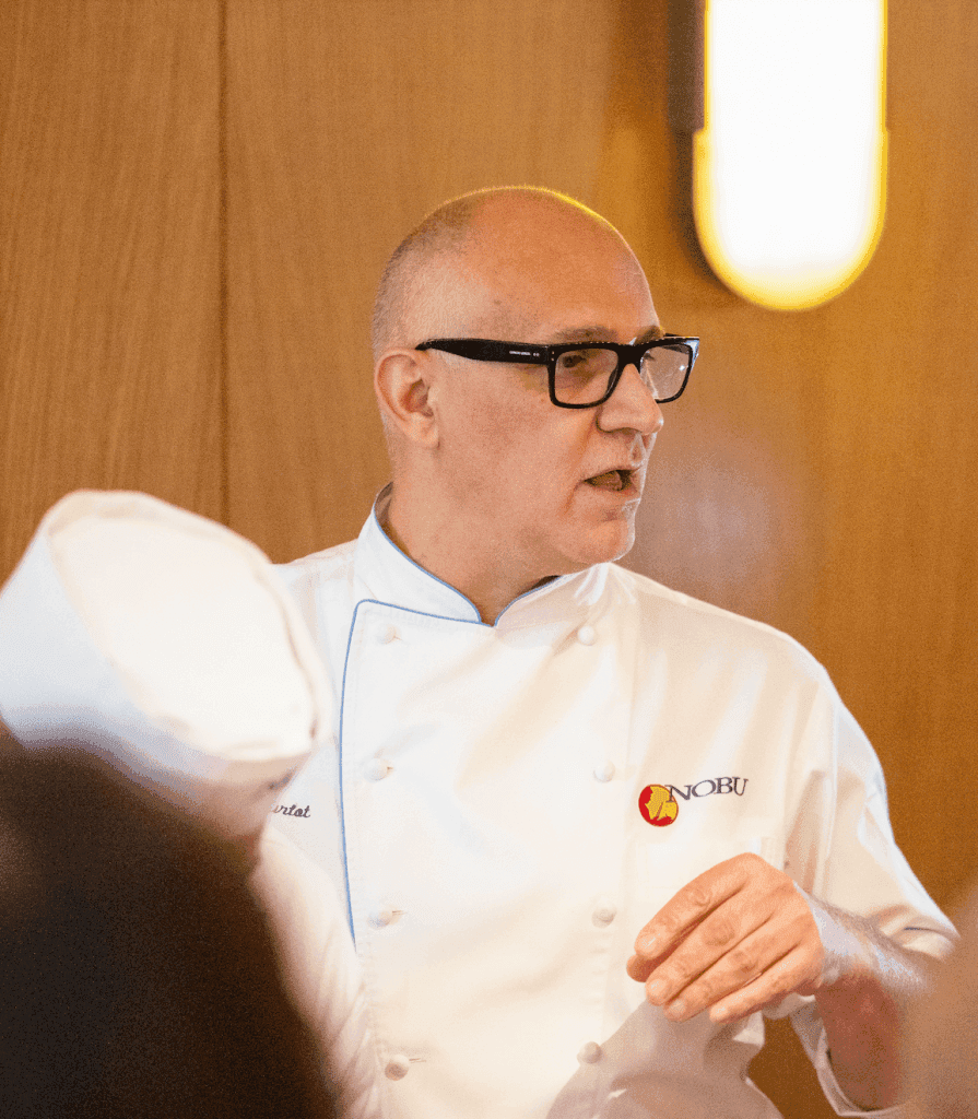 Chef Hervé Courtot speaking at an event, wearing a white chef's coat with the Nobu logo, gesturing with his hand, and appearing to share insights about the restaurant's growth and philosophy.