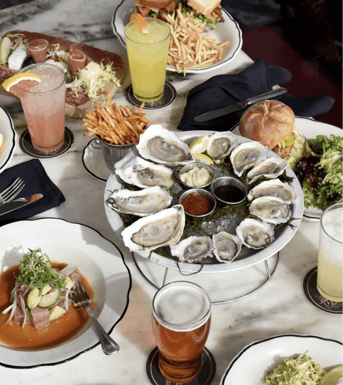 A table set with a platter of raw oysters, fries, salads, sandwiches, various drinks, and dipping sauces on white plates.