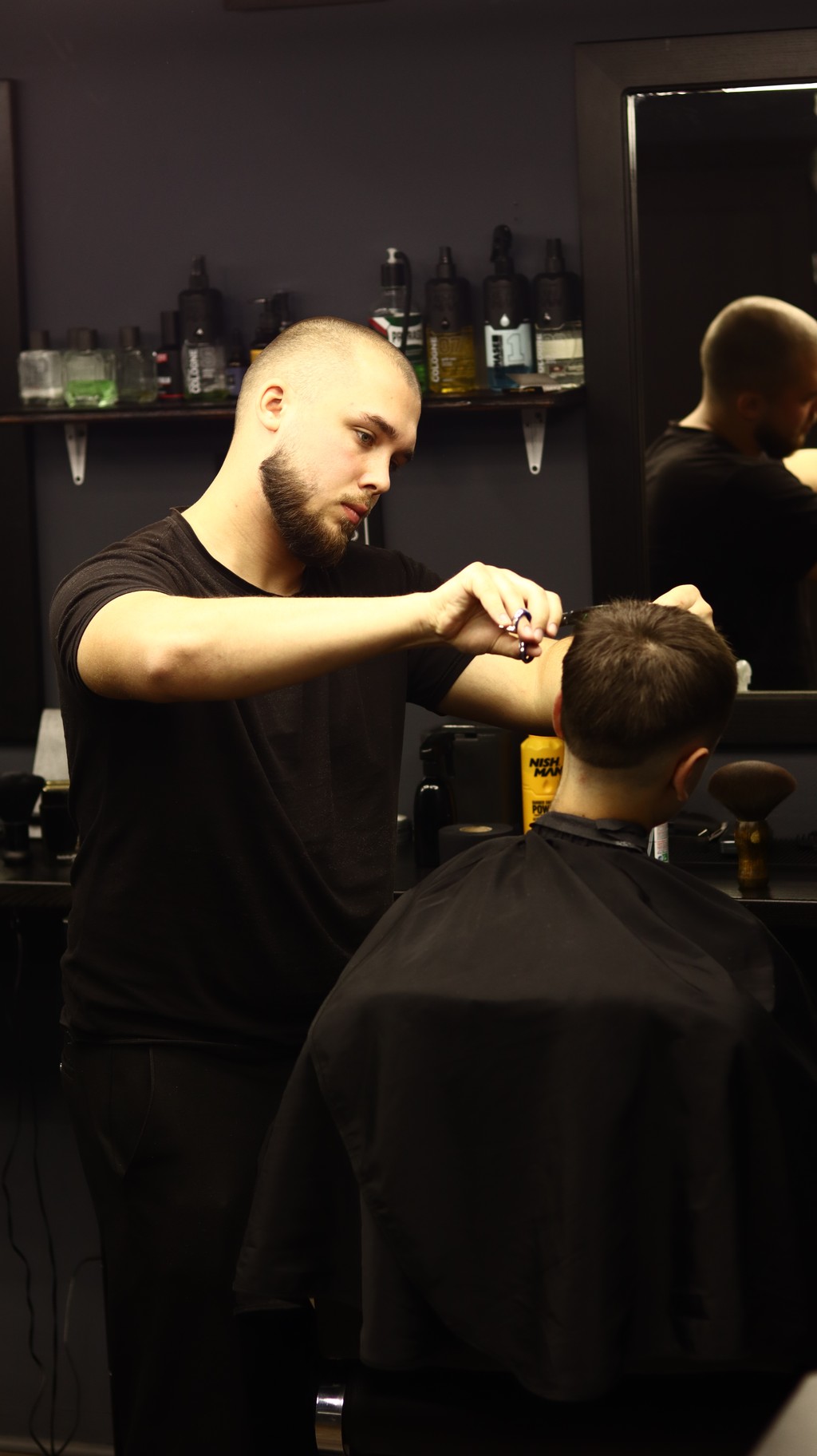 A barber styles a client's hair in a modern barber shop, with tools and mirrors in the background.