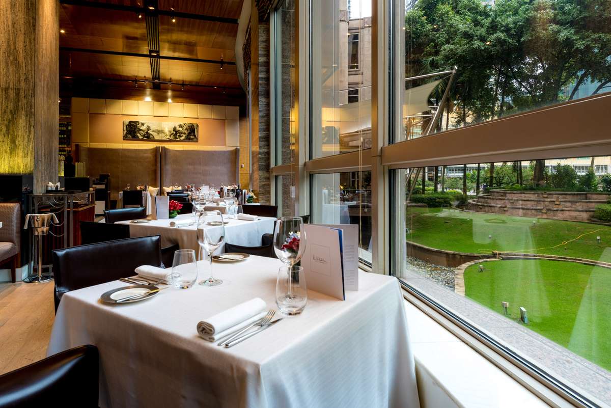 A formal dining room with white tablecloths and leather chairs overlooks a large window facing a green lawn and trees.