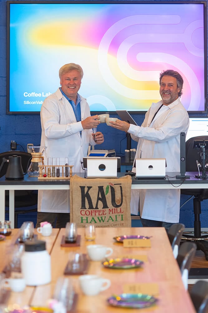 Two individuals in lab coats exchange an item, smiling, in front of a colorful backdrop. A table is set with various items.