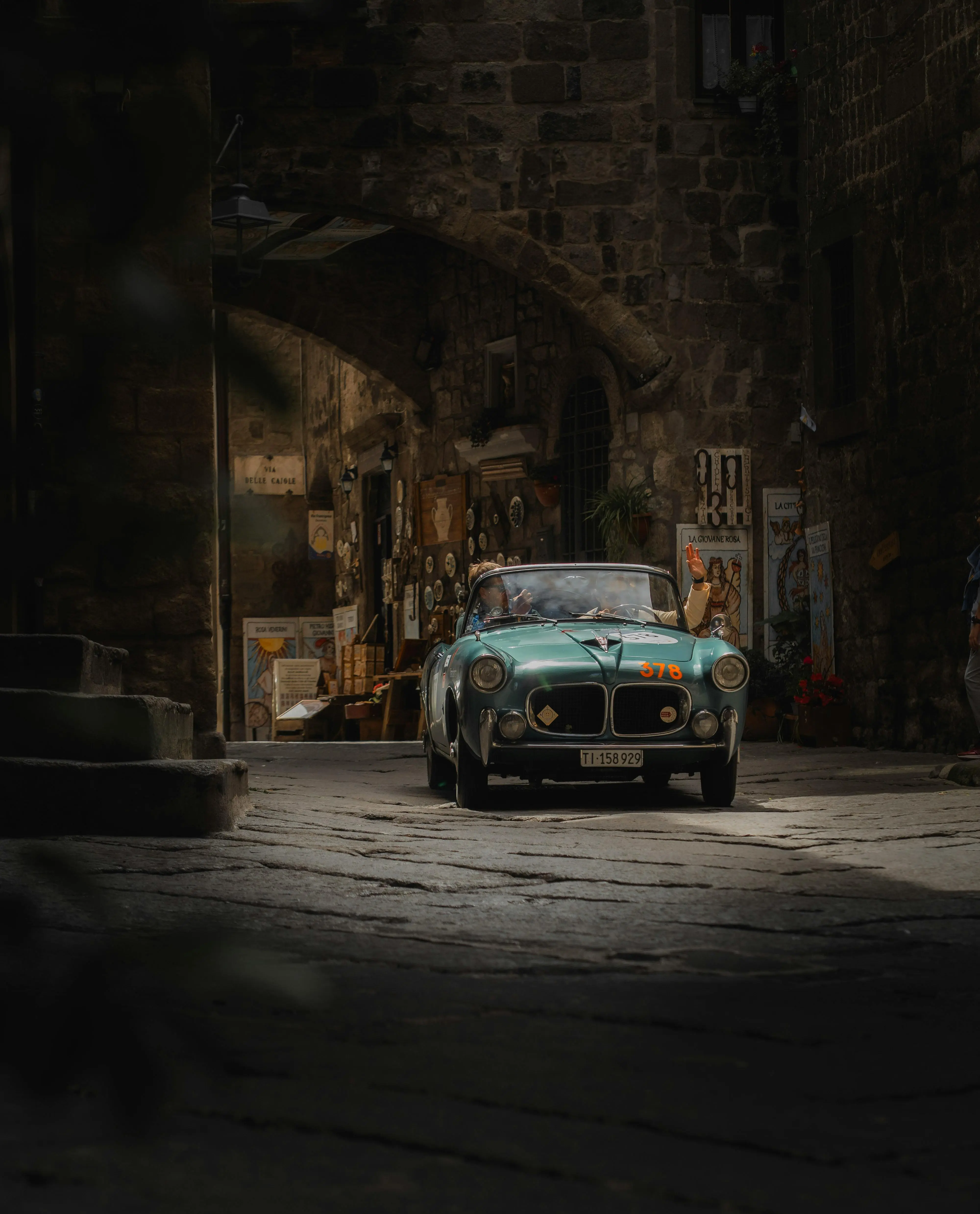 A vintage classic car, a green 1950s model, drives through a narrow cobblestone street in a charming, old town with stone buildings and retro signage, evoking a nostalgic, timeless atmosphere.