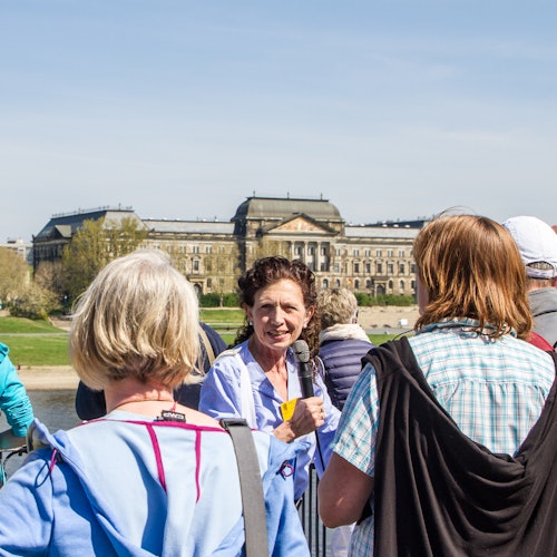 Opas puhuu pienelle ryhmälle ihmisiä, suuren rakennuksen ja nurmialueen ollessa taustalla kirkkaan sinisen taivaan alla.