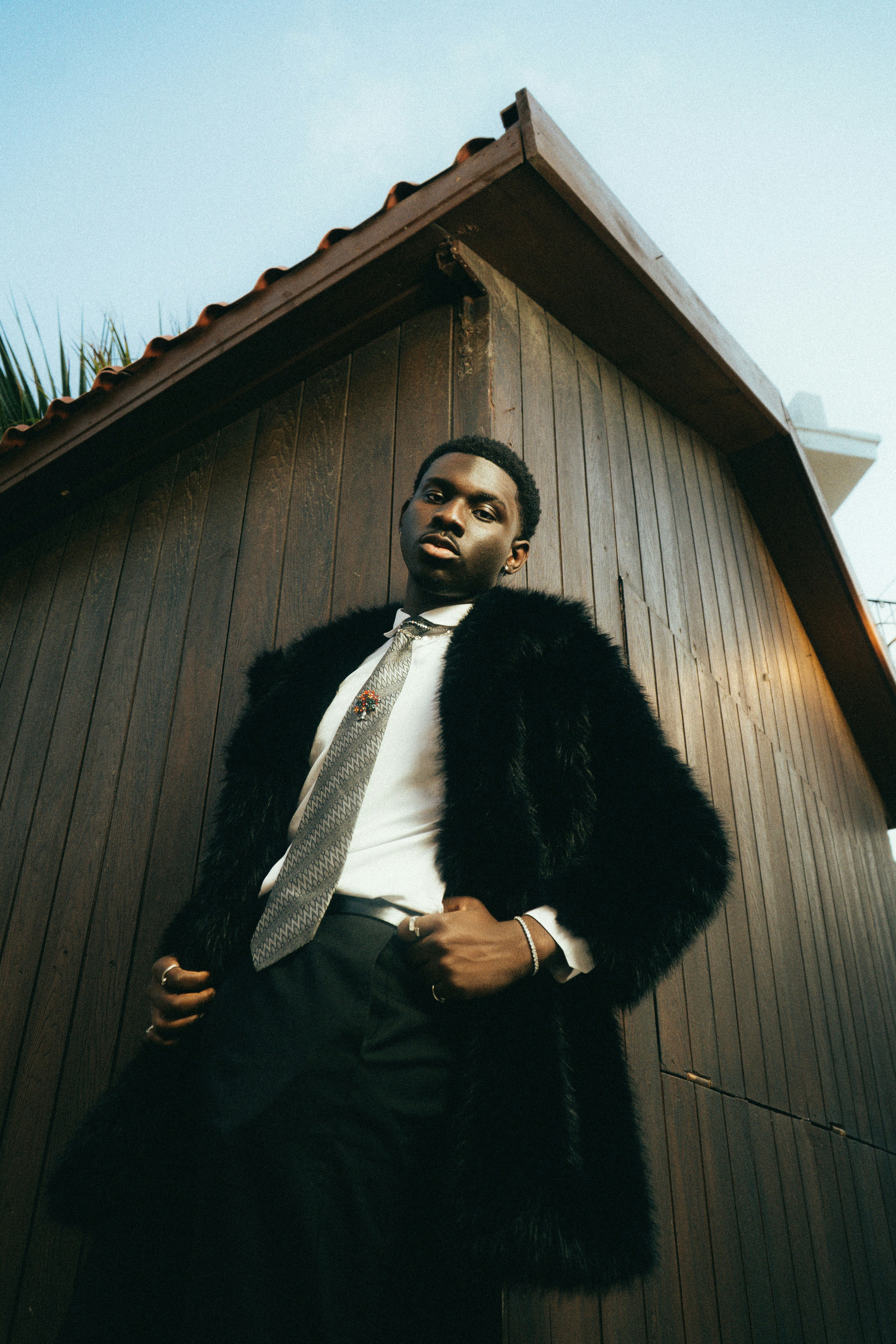 Man in fur coat and tie leans against wooden wall