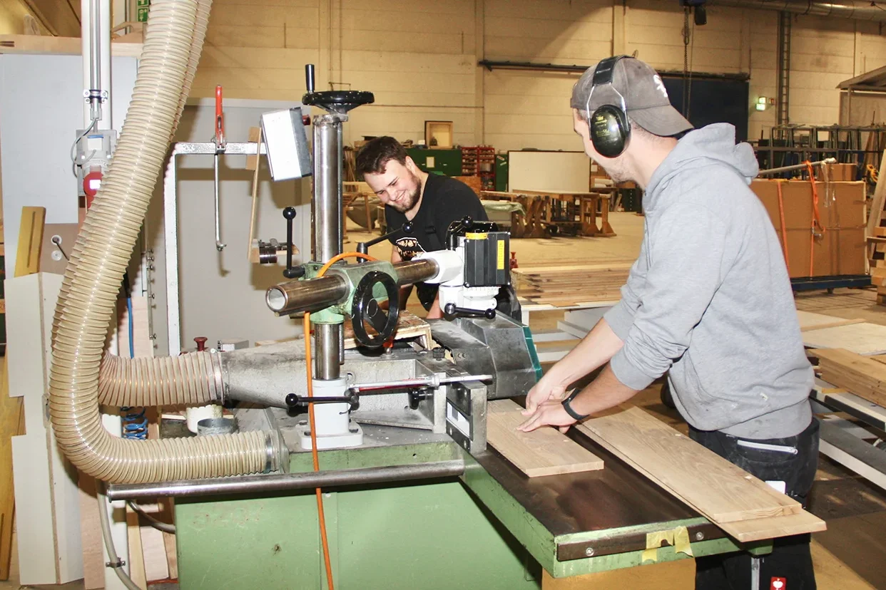 Zwei Männer arbeiten mit einer Hobelmaschine in einer Werkstatt, beide in Arbeitskleidung und mit Gehörschutz.