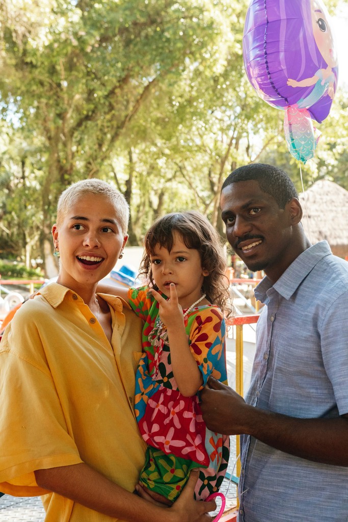 A happy family enjoying a day outdoors, with a mother holding her young daughter in a colorful dress and a father standing beside them, all smiling warmly, accompanied by a vibrant balloon in the background.