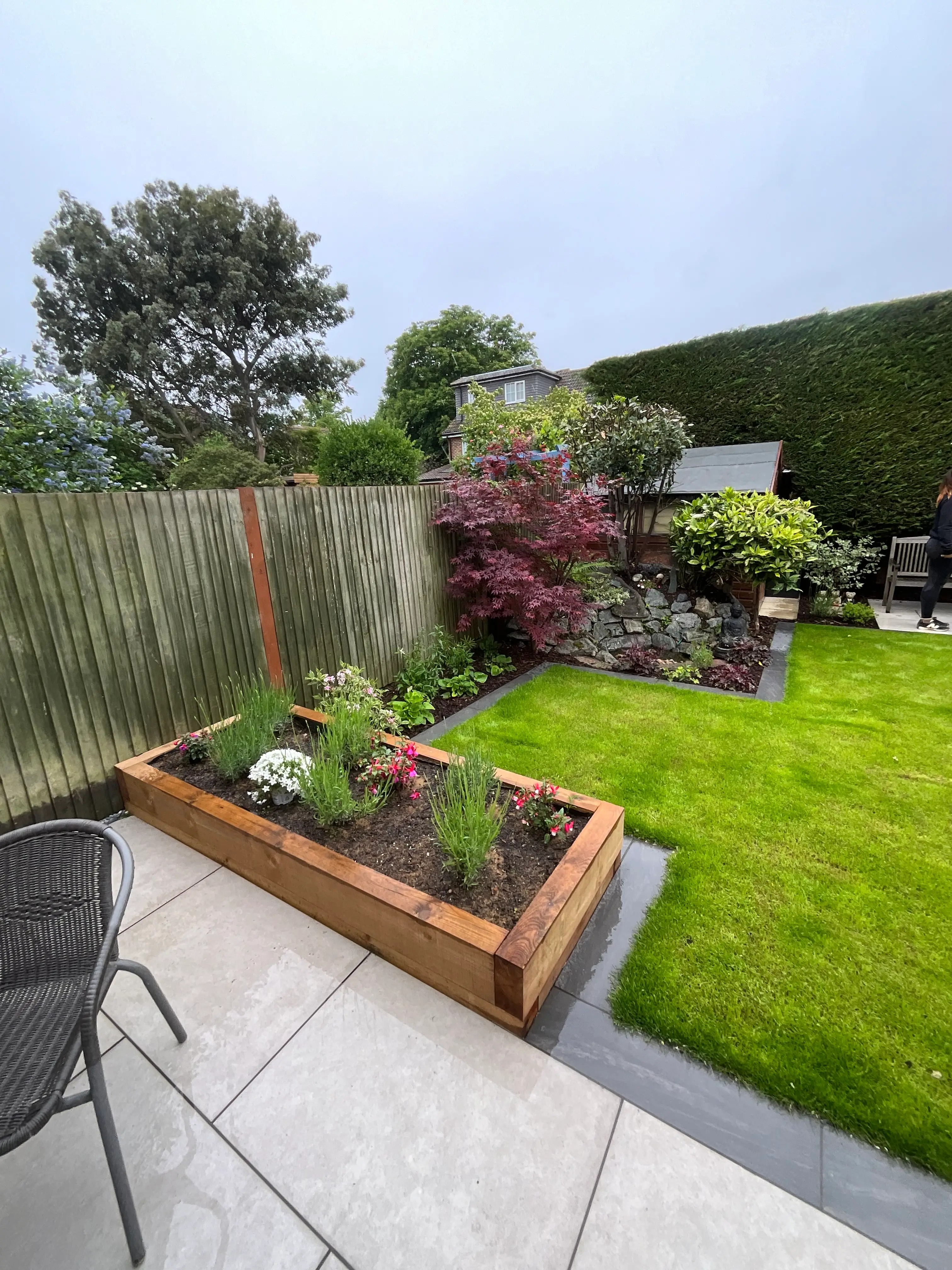 A well-maintained garden featuring a grassy area, a wooden planter, and various plants against a cloudy sky.