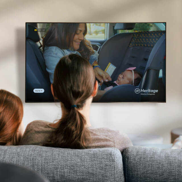 Two people sit on a couch watching a TV screen displaying a woman buckling a baby into a car seat.
