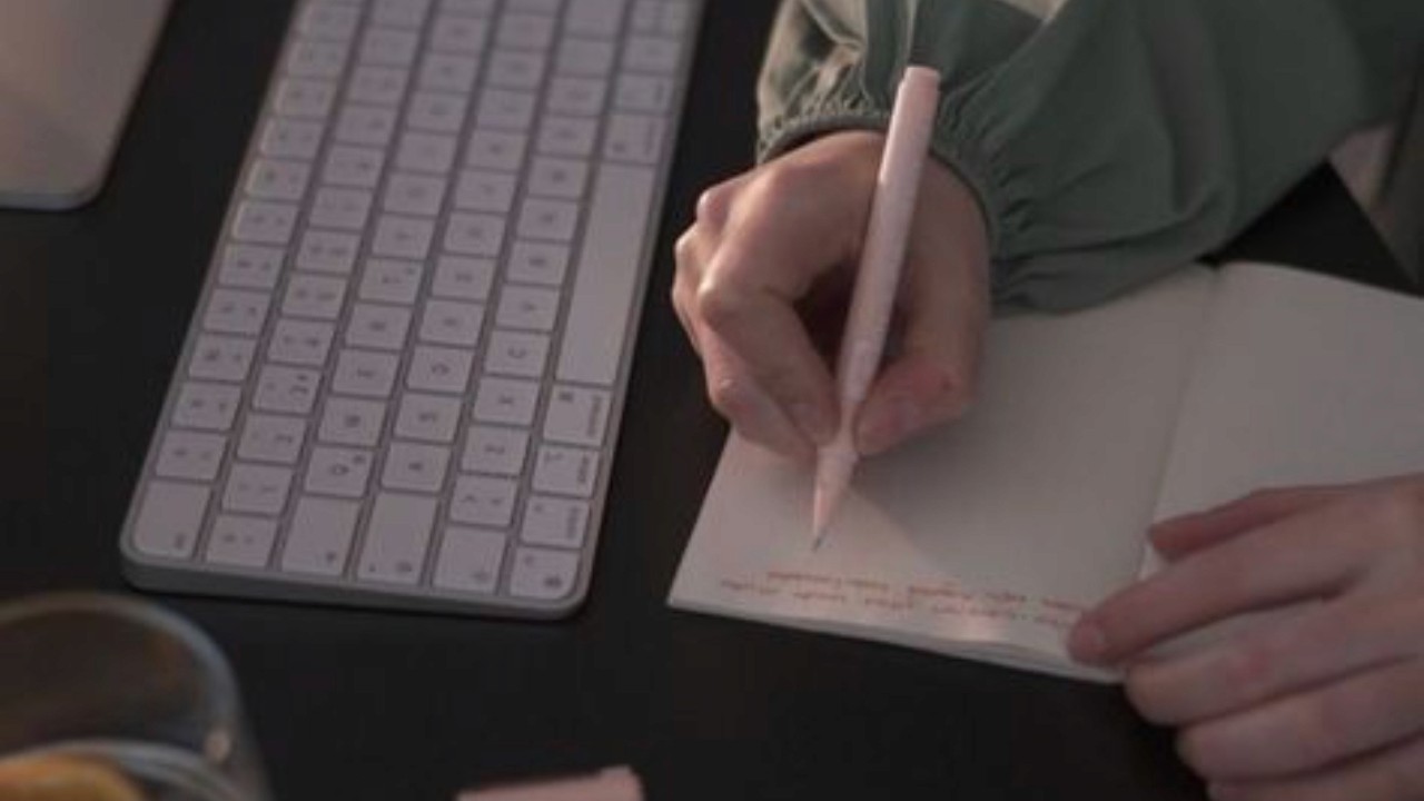An image showing a hand writing in a notepad with a pen and a keyboard on the side