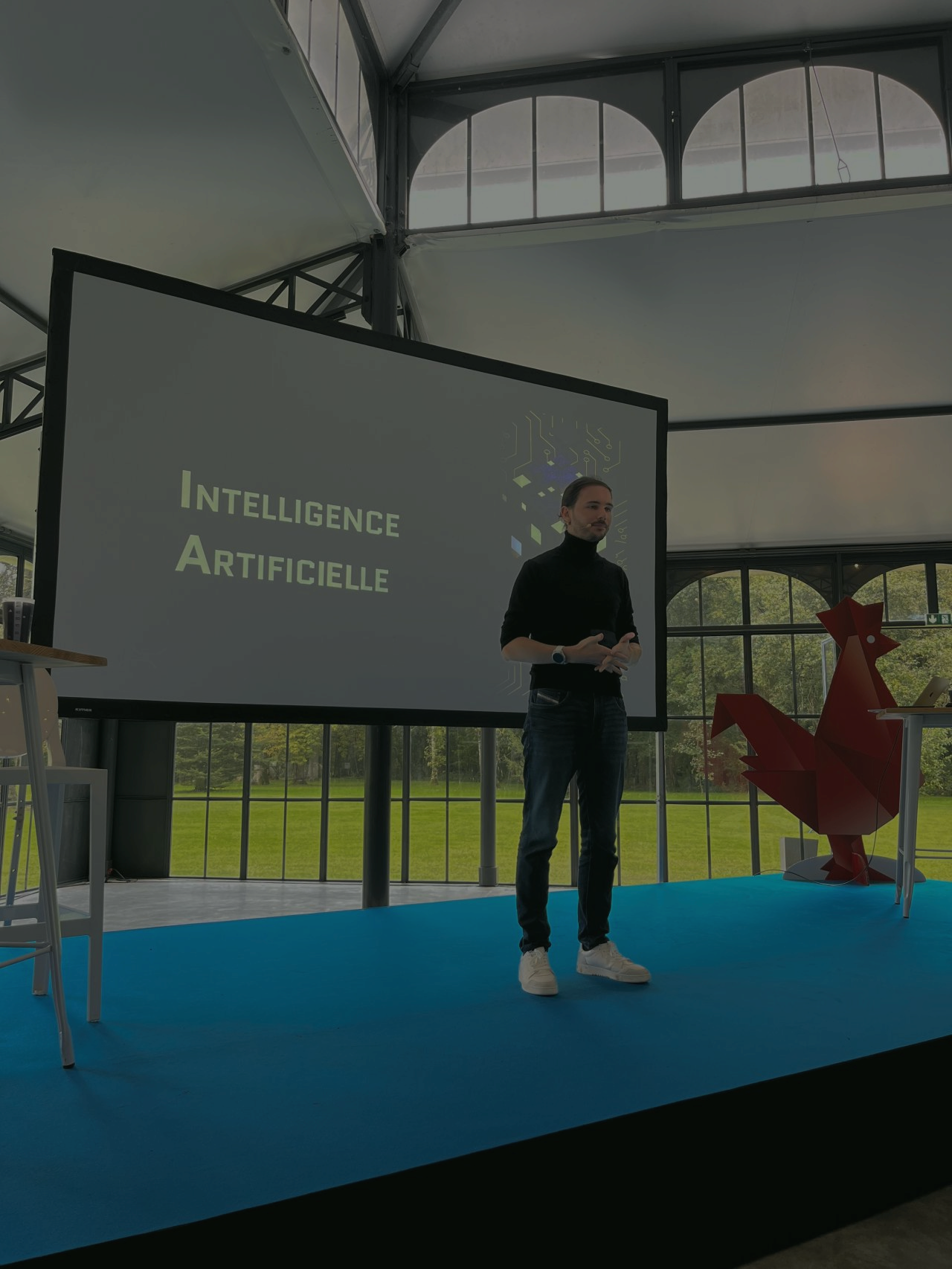 A person stands against a vibrant background of orange and yellow wavy patterns. They are wearing a dark coat, looking contemplative and thoughtful.