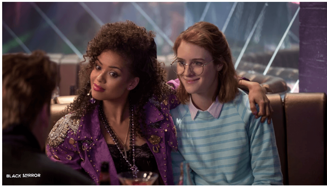 Two main characters from the Black Mirror episode 'San Junipero,' sitting together in a retro-style bar, one with curly hair wearing a purple jacket and the other with glasses and a striped shirt, smiling and embracing.