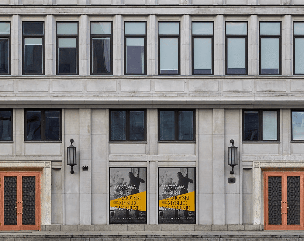 A symmetrical architectural photograph of a modern stone building featuring two August Zamoyski exhibition posters at the entrance. This highlights the studio's talent for creating consistent brand applications across diverse architectural contexts.