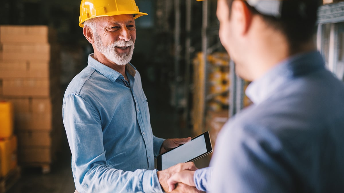 Two men shake hands in a warehouse; one is wearing a yellow hard hat and safety gear.