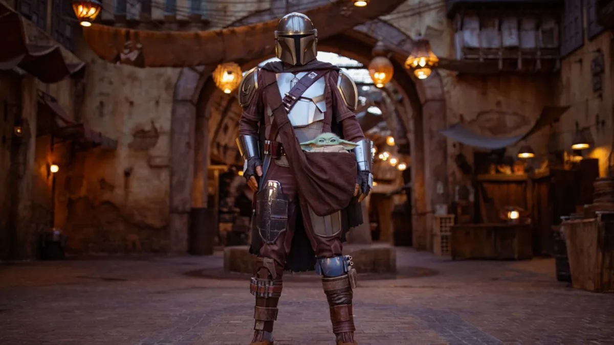 The Mandalorian, clad in his signature beskar armor, stands tall in the bustling streets of Batuu. Grogu peeks out from his satchel, curiously observing the world of Star Wars: Galaxy’s Edge.