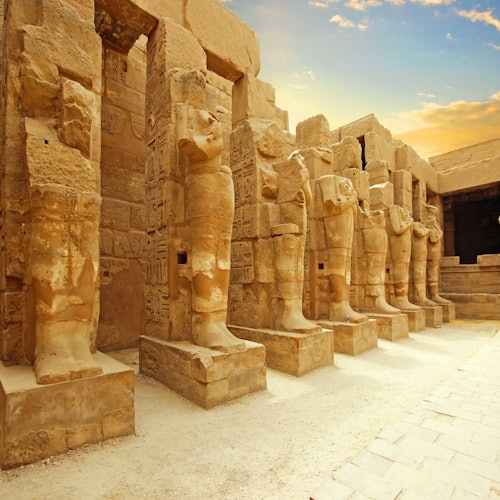 Ancient stone statues stand in rows under a partly cloudy sky at an Egyptian temple site.