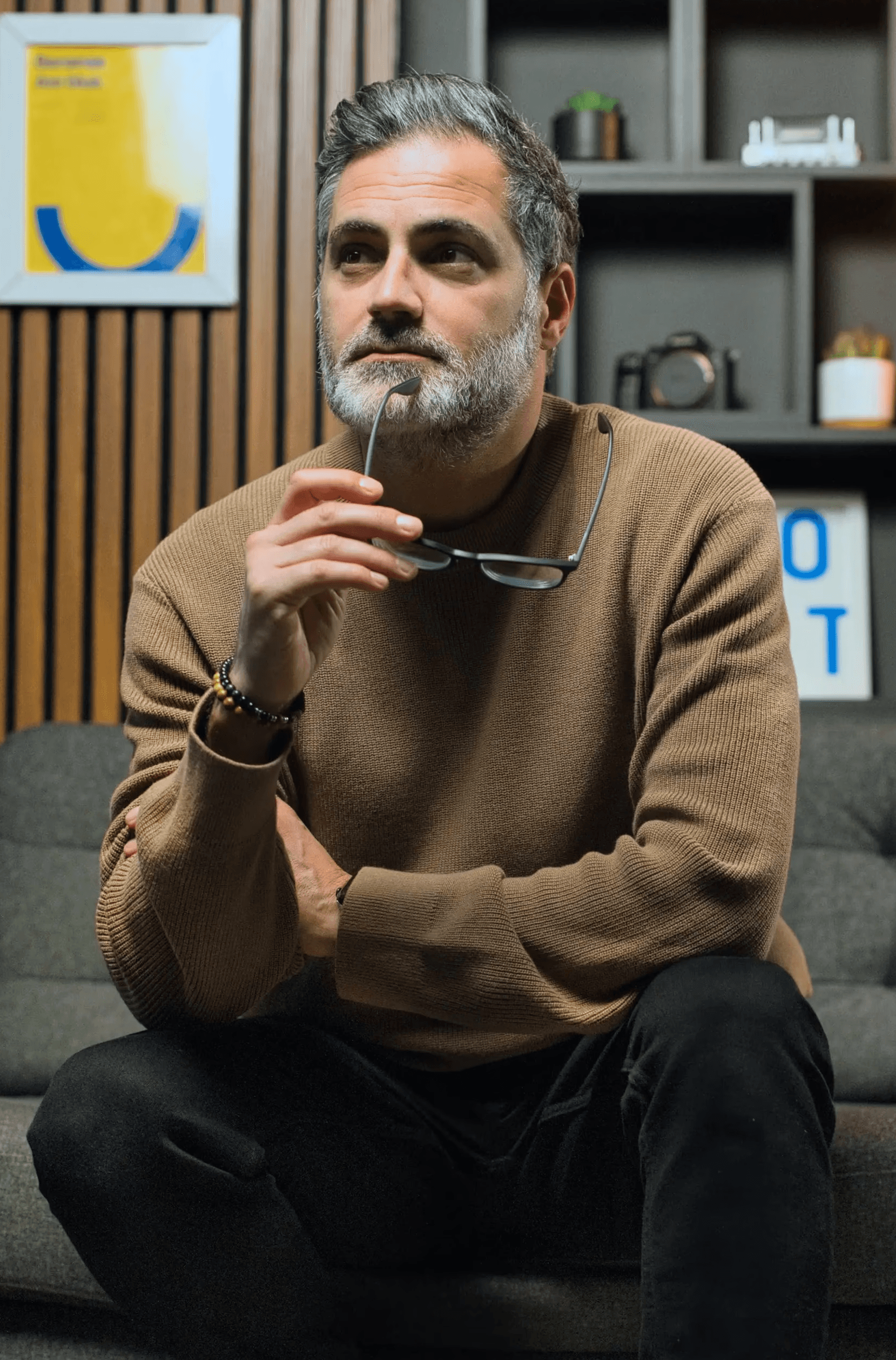 A thoughtful individual with a beard sits on a gray sofa, holding glasses in one hand, against a backdrop of wooden paneling and minimalistic shelves adorned with various decorative items.