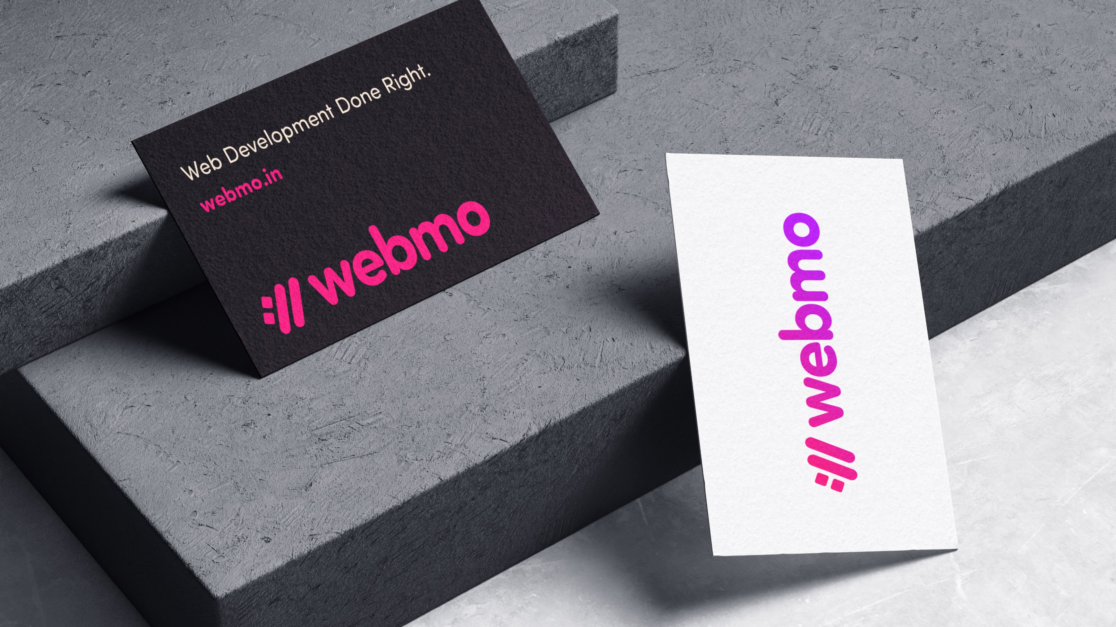 A mock-up of a black business card with a brand identity design, magenta logo at the bottom, big and bold text on the top, and another business card front design that has the Webmo logo in a gradient from pink to purple. The business cards are sitting on a marble modern architecture tabletop. 