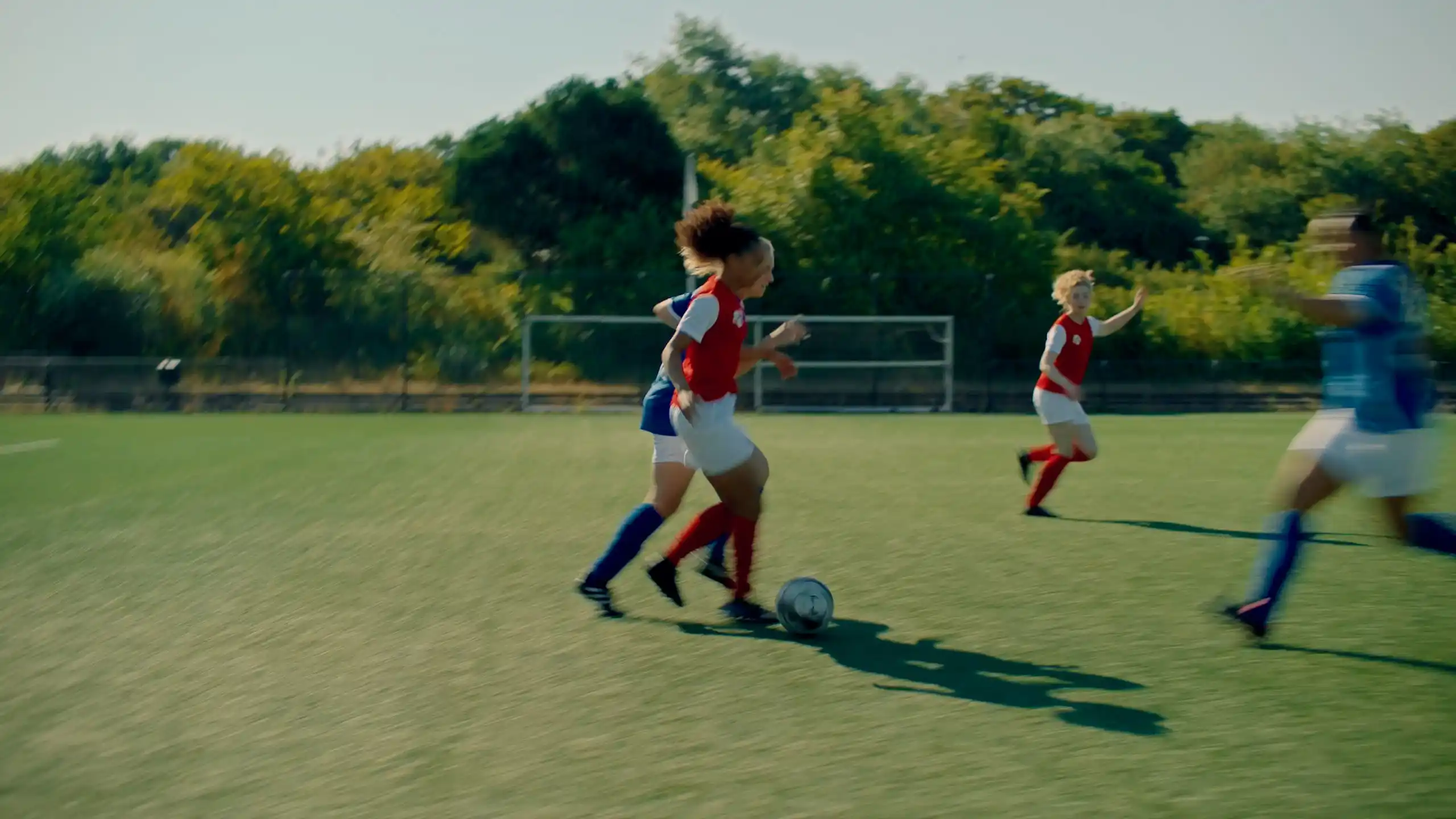 Wide shot of female football players chasing the ball across a sunlit pitch.