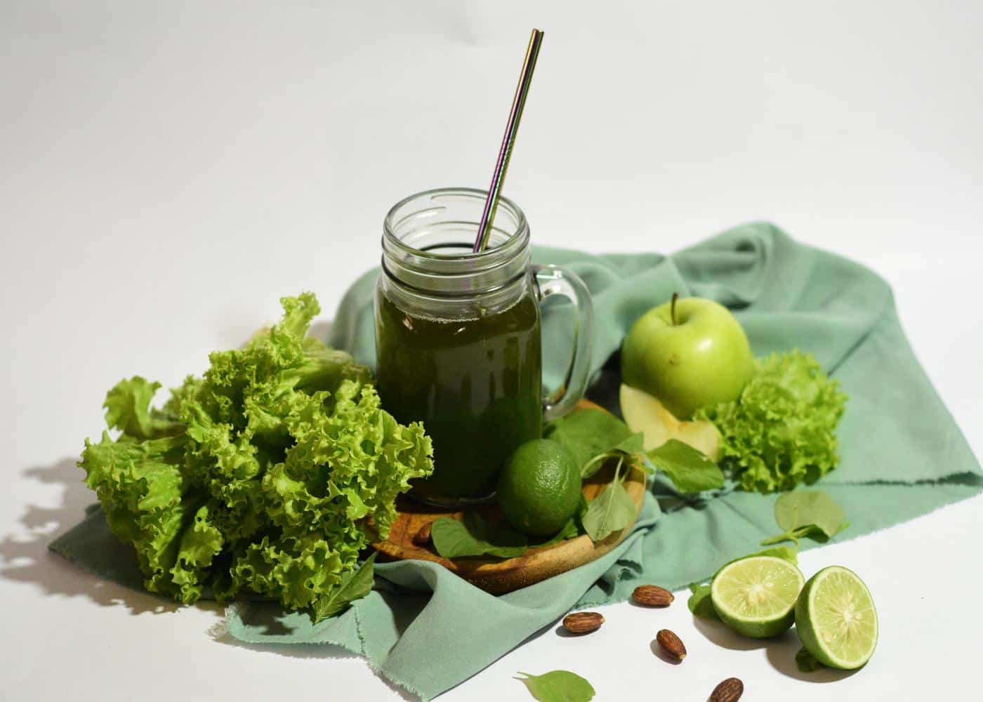 Mason jat with green juice next to kale, limes, and green apples on top of a green cloth