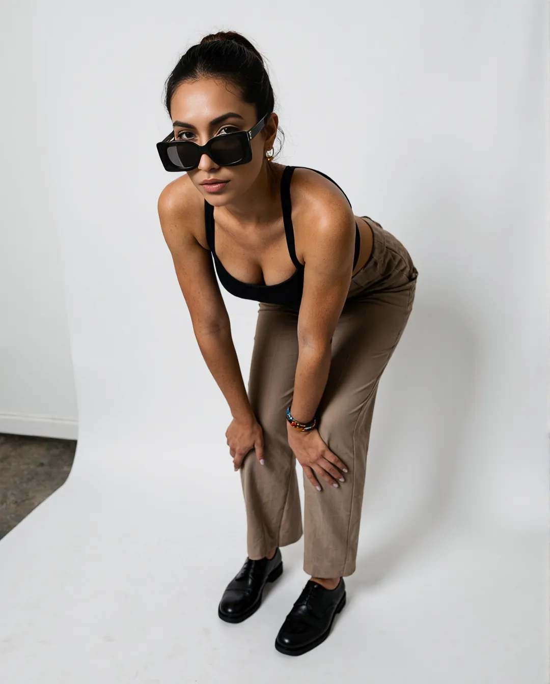 Minimalist fashion portrait featuring oversized sunglasses, black tank top, tan trousers and black shoes against a clean white backdrop