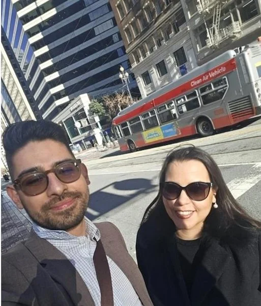 Um homem e uma mulher, ambos sorrindo, estão em uma rua urbana ensolarada, com edifícios modernos altos e um ônibus urbano vermelho ao fundo.