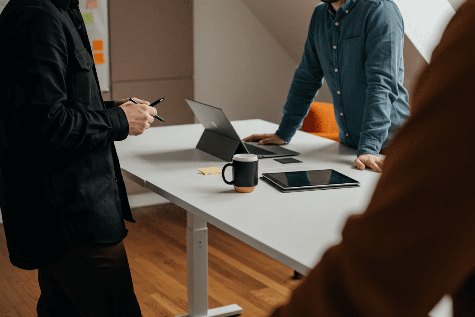 Group of people standing around a white desk during a casual meeting with laptops and coffee cups.