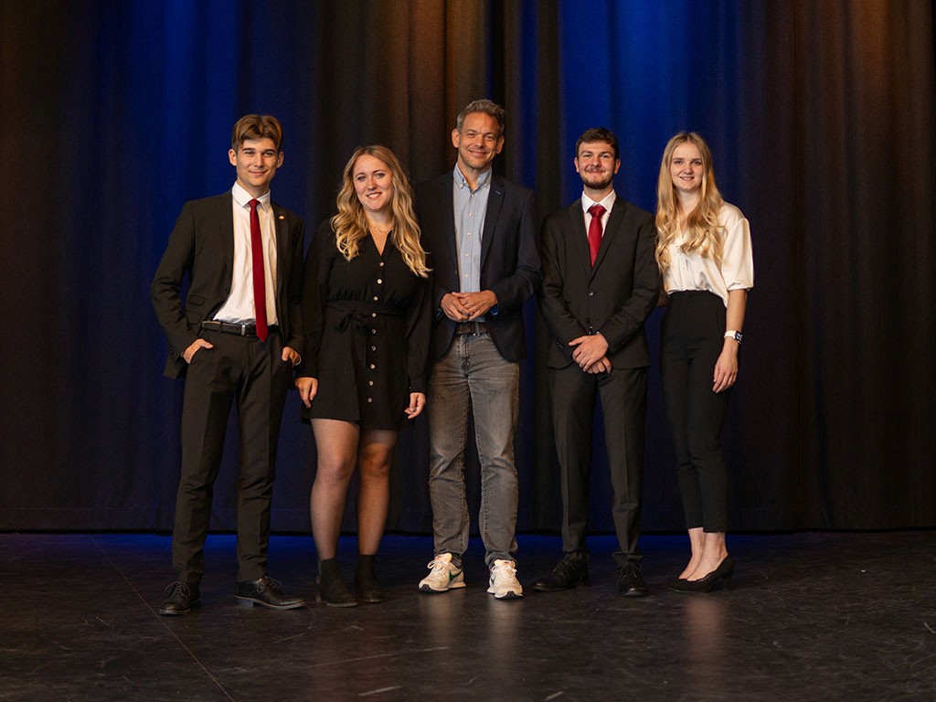 Debattenschmiede-Team präsentiert sich im Business-Outfit auf der Bühne nach erfolgreicher Jugenddebatte.