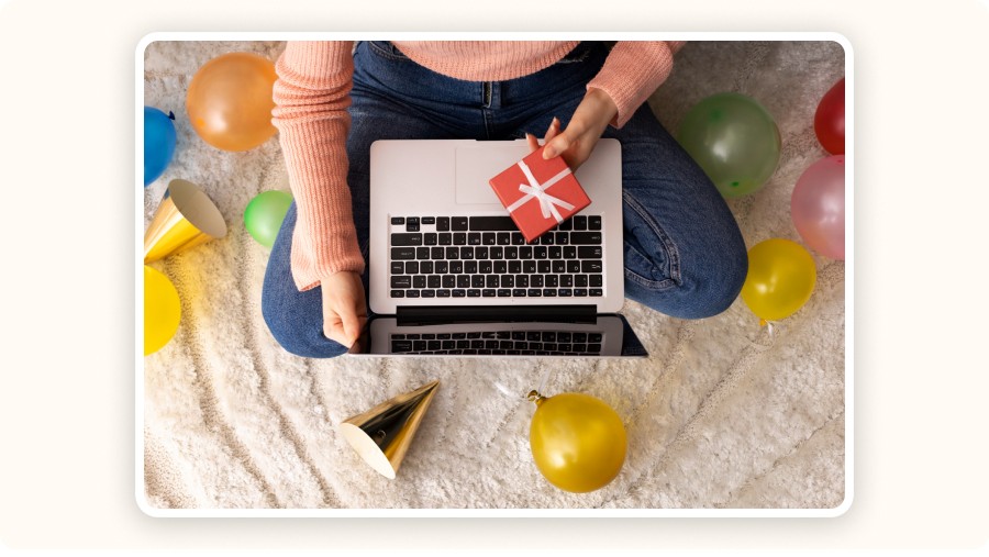 Person holding gift while shopping online, surrounded by colorful balloons.