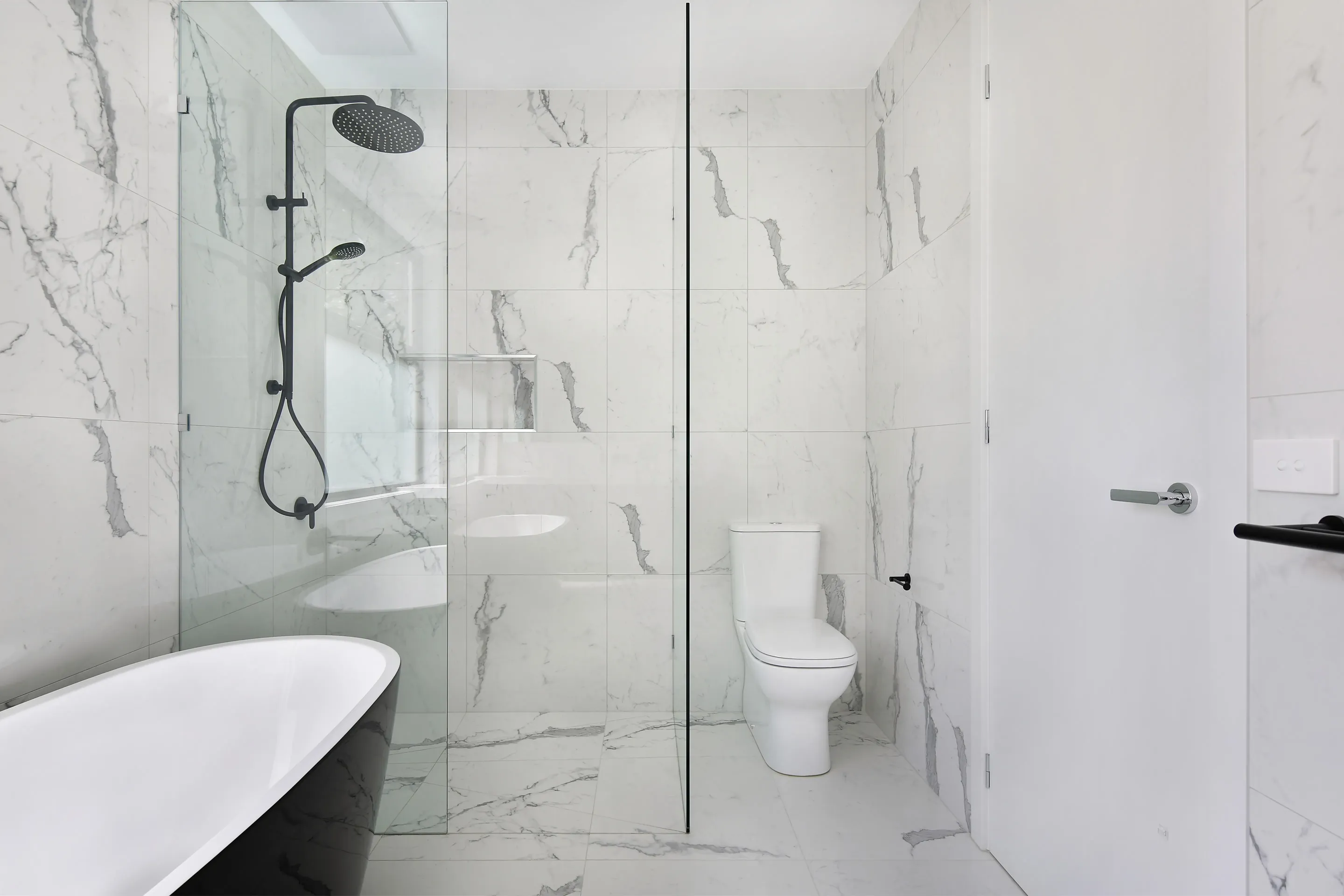Marble-tiled bathroom with a glass-screened shower with black rail and rain head, a recessed niche, a wall-hung toilet and a white door with chrome lever