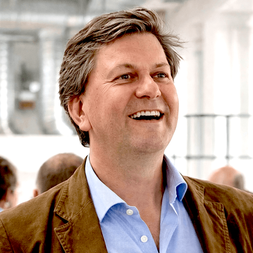 A smiling man in a suit and glasses sits at a table, with a modern office backdrop.