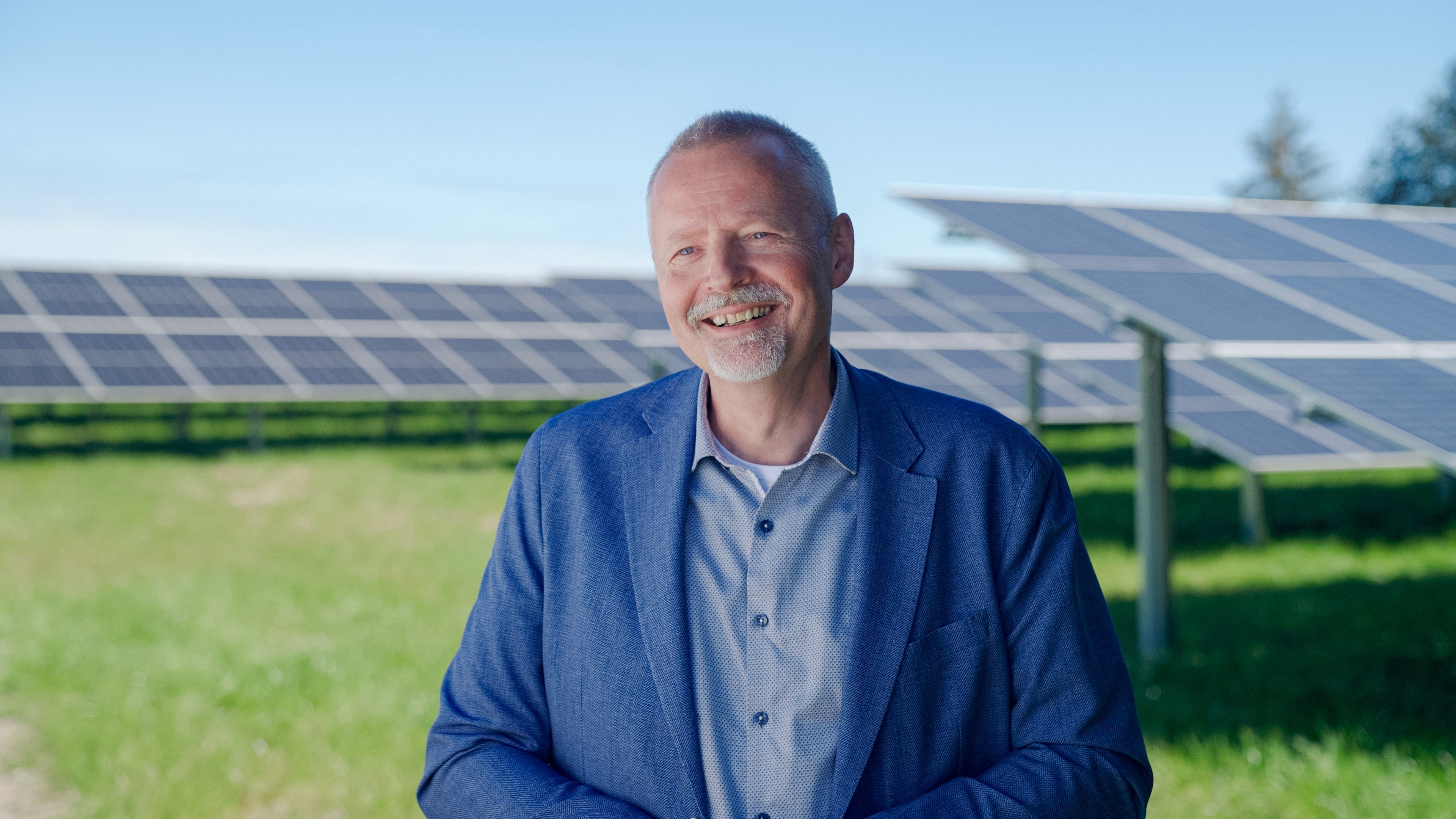 Man with white beard and wearing blue suit, smiling and standing in front of a field of solar panels.