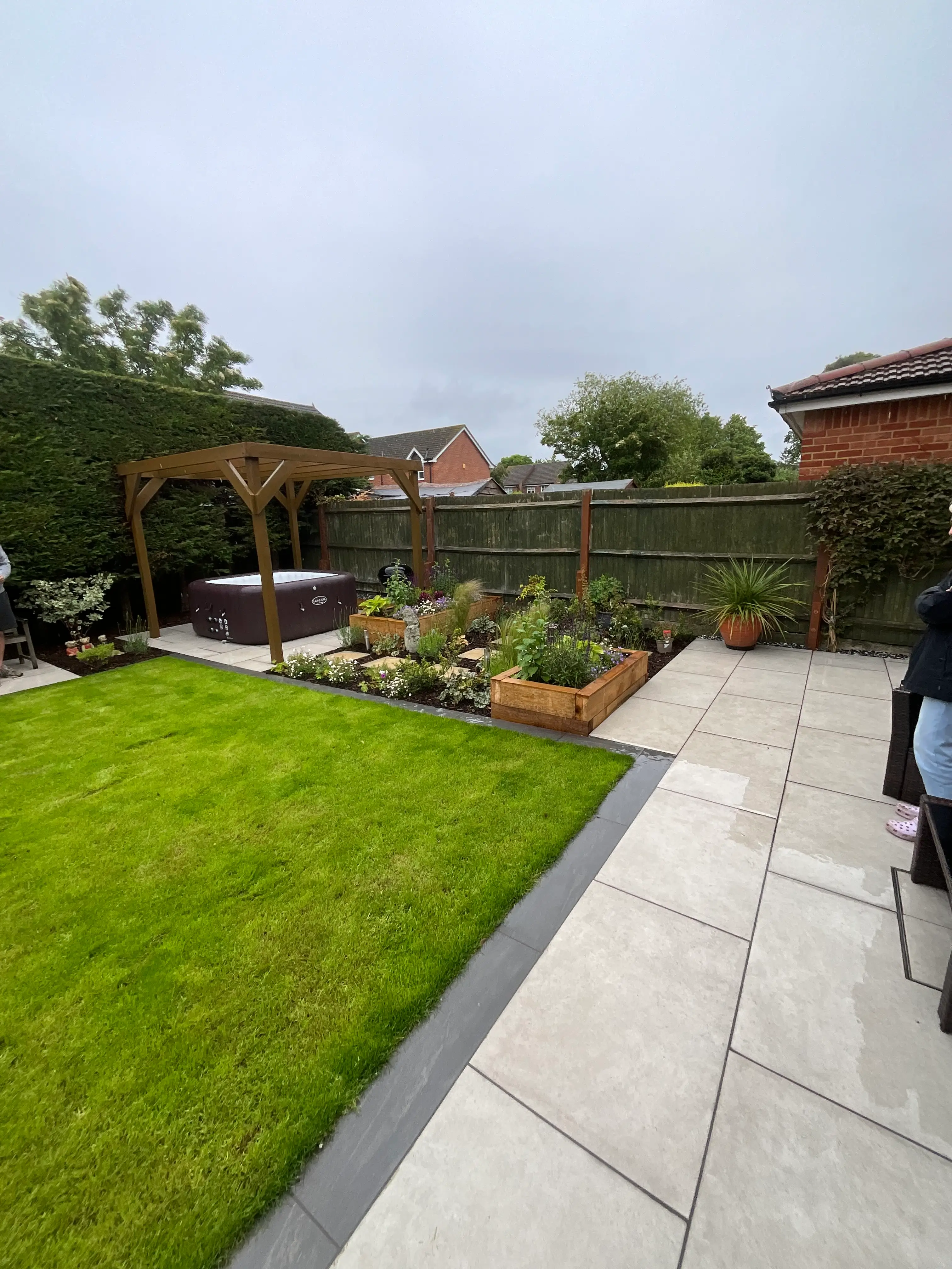 A view of a garden with green grass, plants, and houses in the background under a cloudy sky.