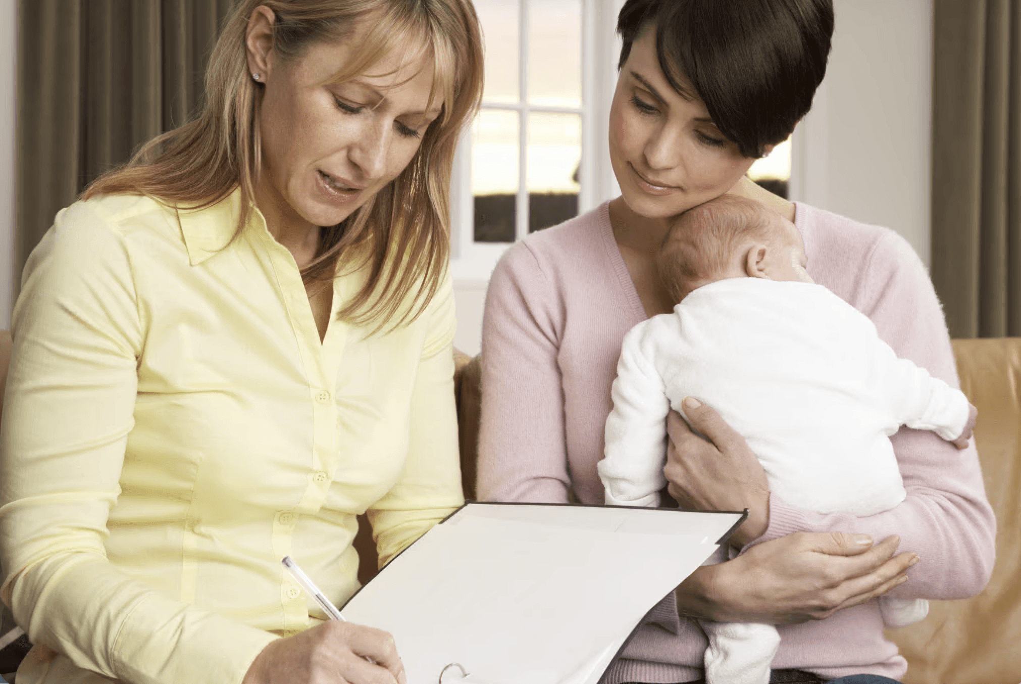woman writing in notebook as second woman holds baby and looks on