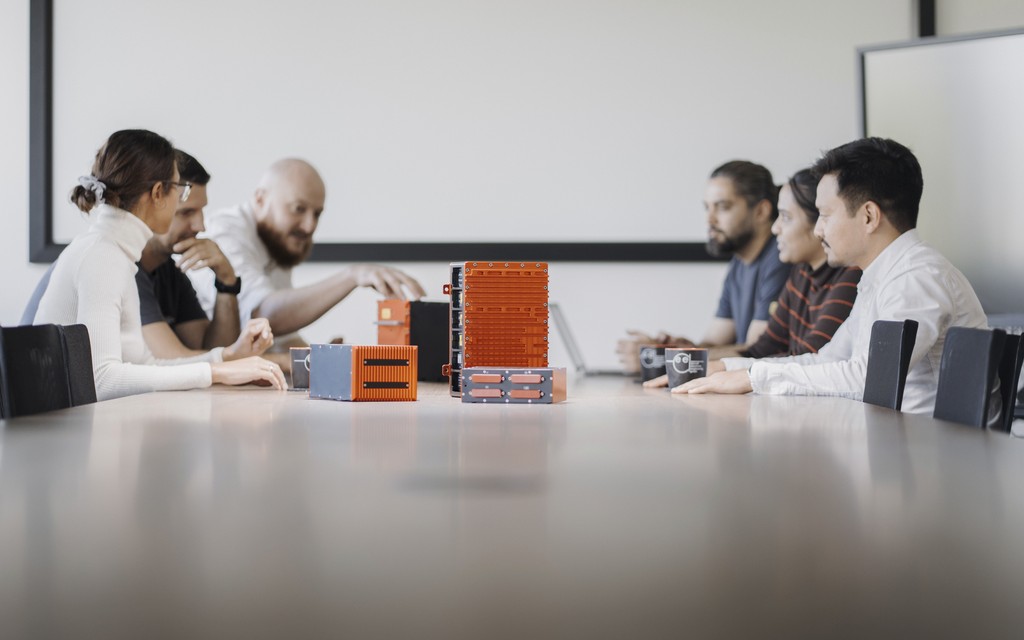 A group of six people engaged in a discussion around a conference table in a bright meeting room. On the table are products of AEE