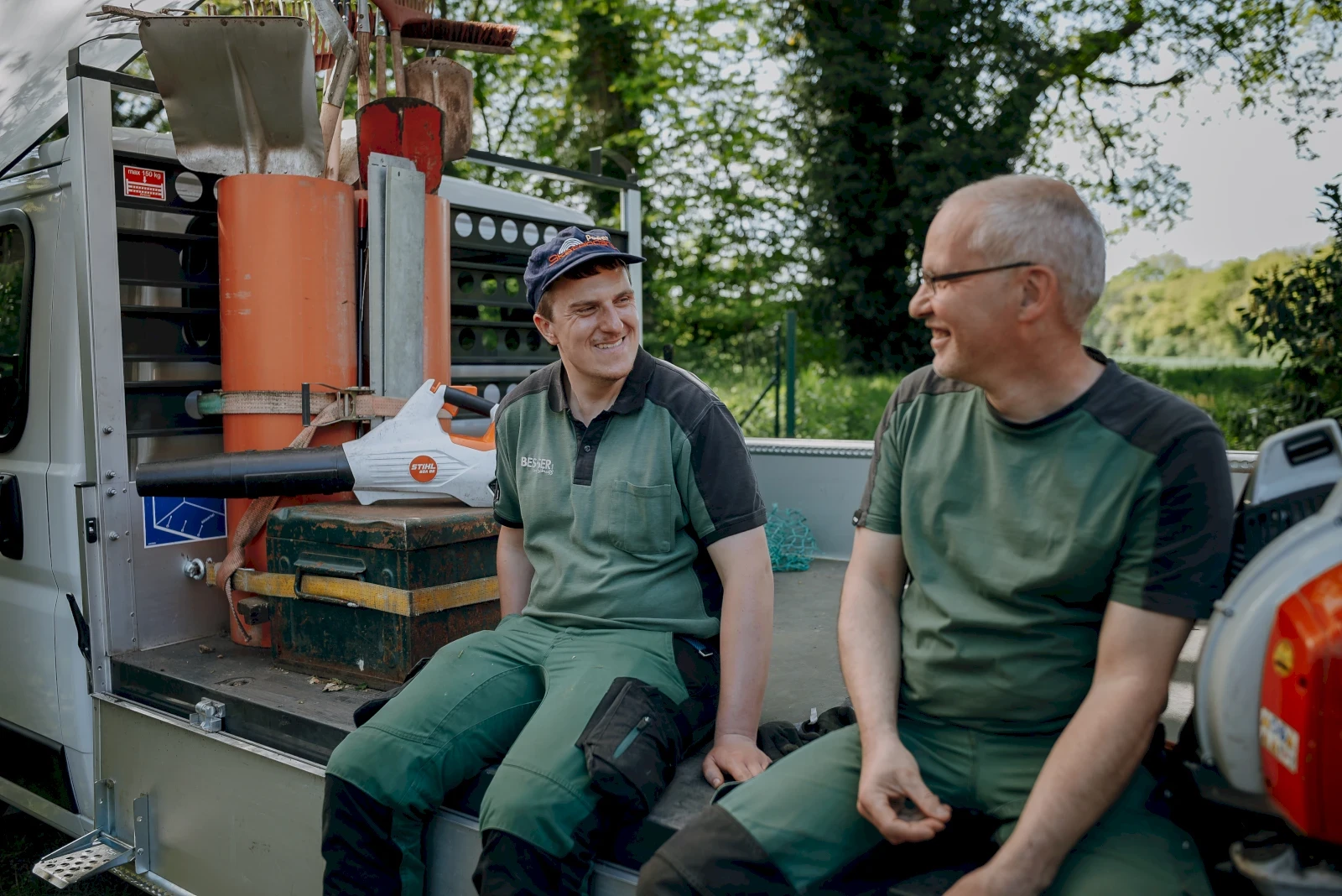 Zwei lächelnde Männer sitzen auf einem Wagen. Beide tragen grüne Arbeitskleidung. Im Hintergrund befindet sich ein Baum.