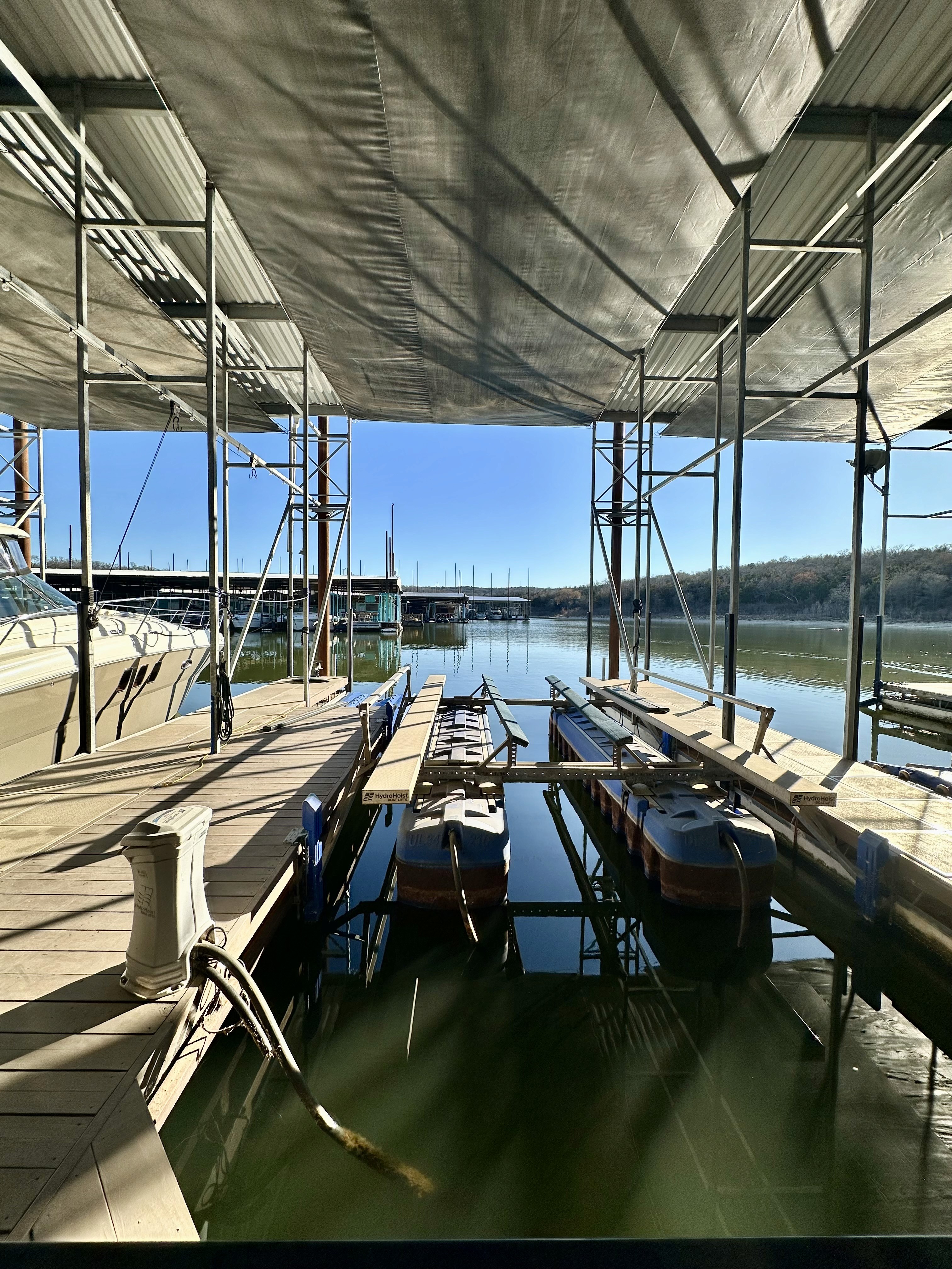 A row of docks sheltered under a metal canopy extends over calm green water, with several small boats moored, against a backdrop of distant trees and clear blue sky.