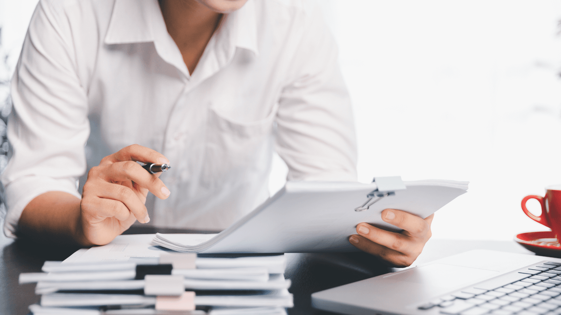 Eine Person in einem weißen Hemd hält ein Tablet, mit einem Stift in der Hand und einer roten Tasse in der Nähe auf einem Schreibtisch.