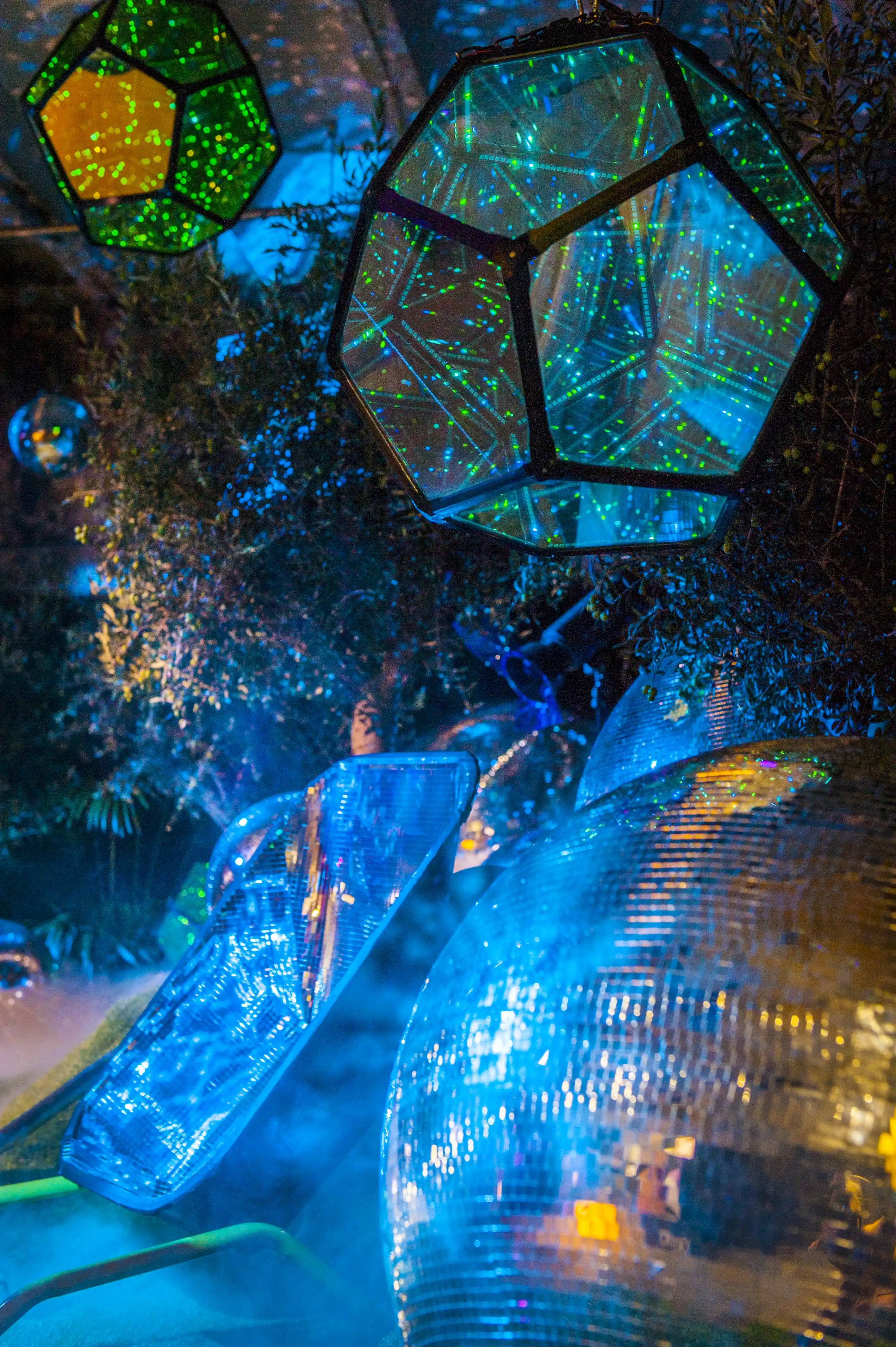 Hanging mirrored dodecahedrons and disco balls glowing green and blue between plants at BERLIN PHOTOWEEK.