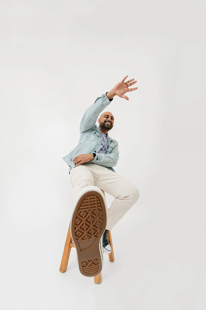 Man smiling sitting on stool wearing denim jacket and cream pants, raising hand with foot extended toward camera against white backdrop.