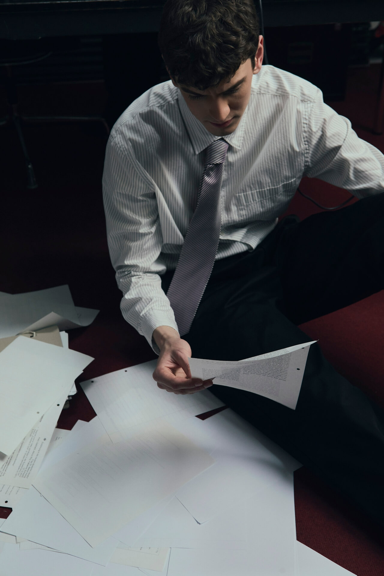 A young man in a white shirt and tie sits on the floor, surrounded by scattered papers. He looks down at a document, appearing focused and pensive.