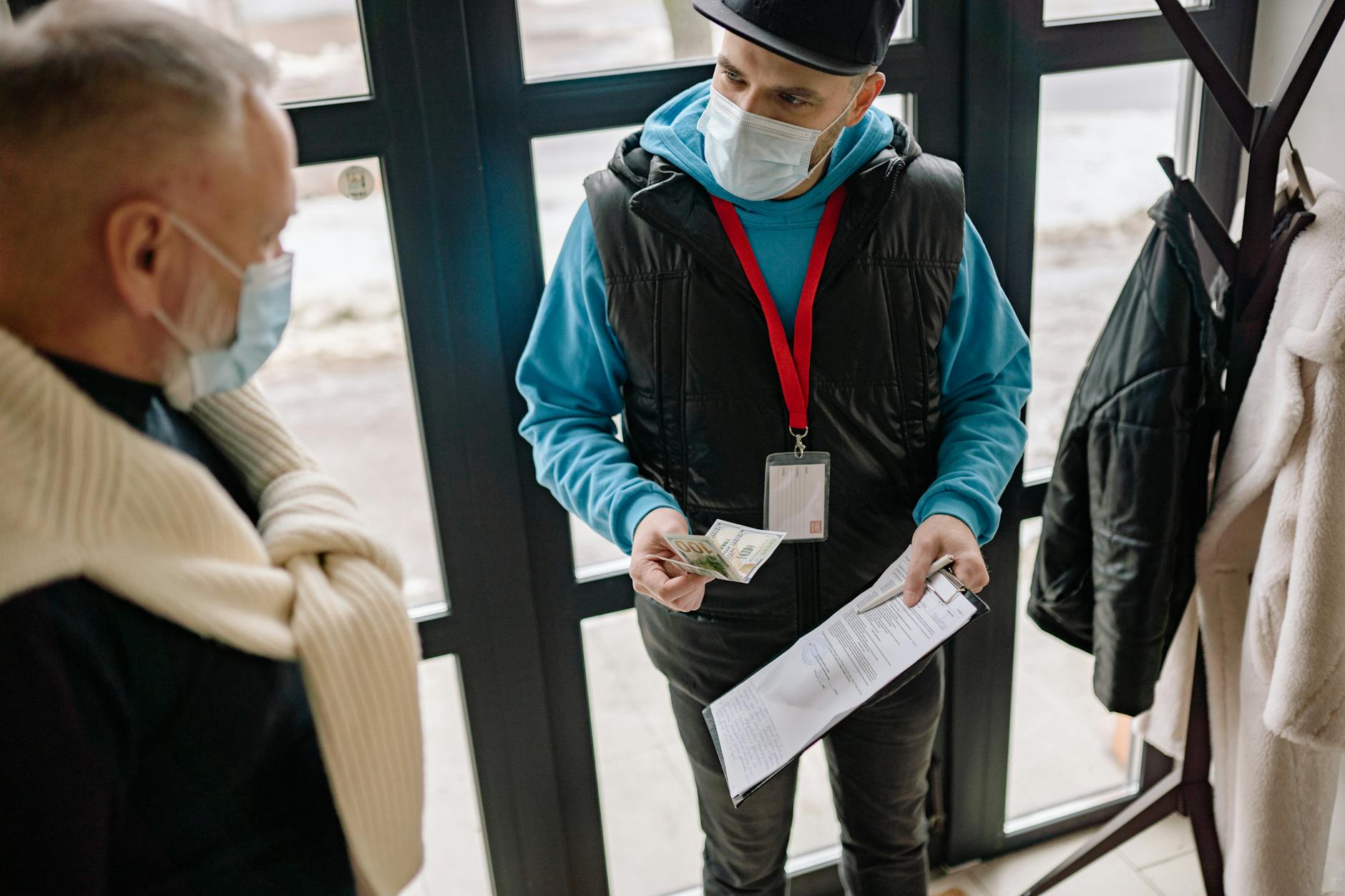 Uniformed official in a face mask counting cash at a front door while presenting documents to a resident
