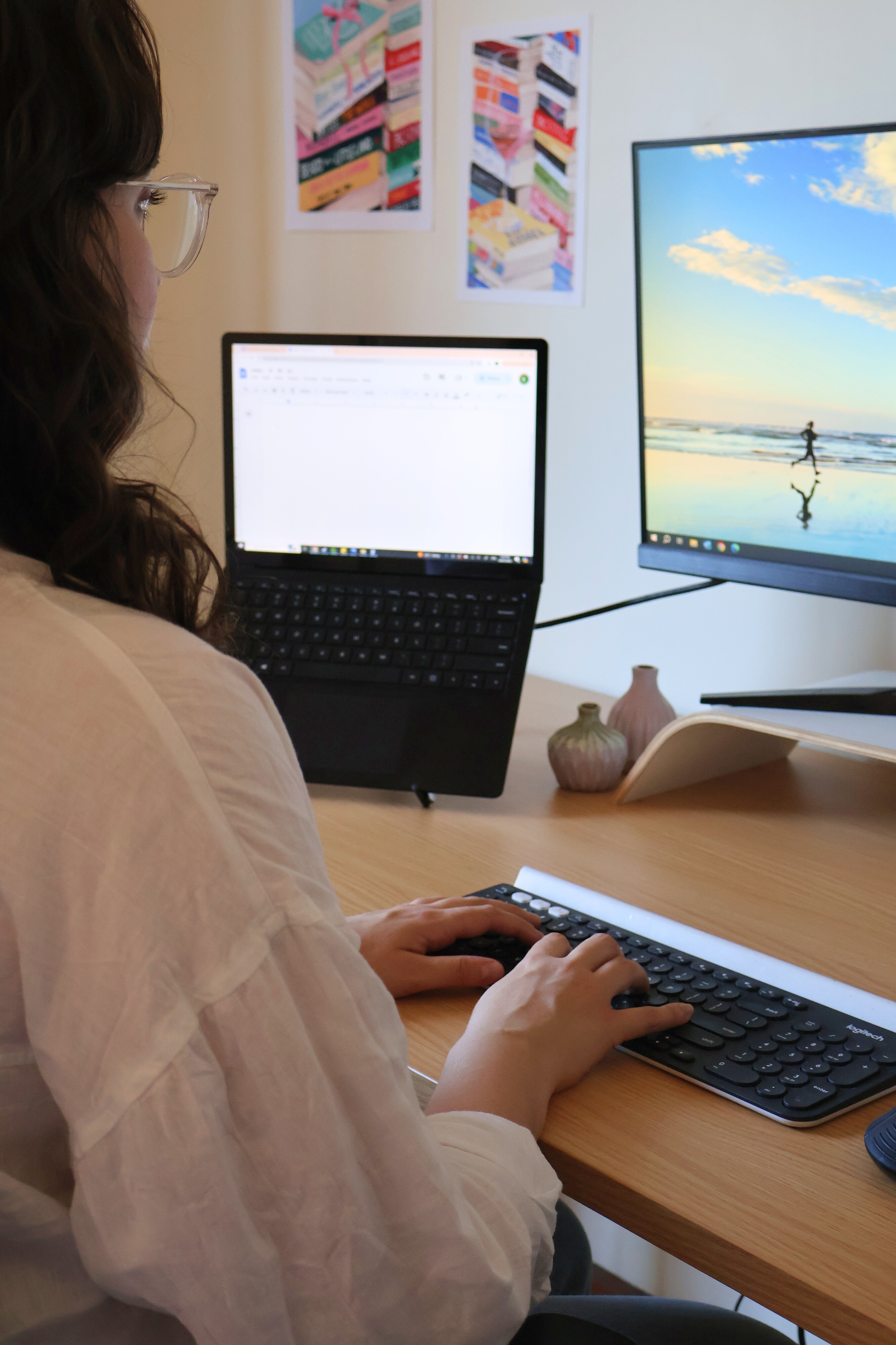 Woman with brown curly hair sitting at a desk working on a desktop monitor. She is typing with both hands on a black and white keyboard. In the background, there are illustrated prints of books on the wall.